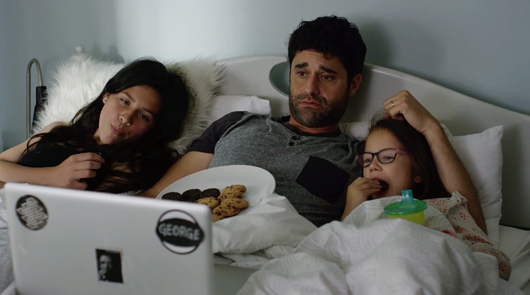 A father and his two young daughters, underneath the covers of a bed. They are watching something on a laptop screen and eating cookies.