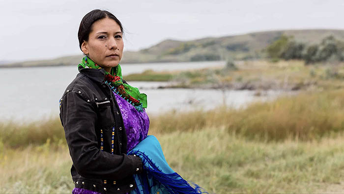 A woman stares at the camera, in the background there is a lake.