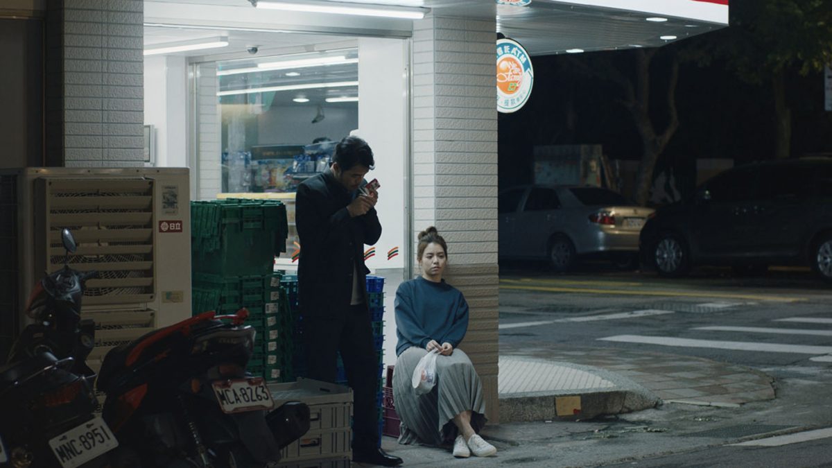A man lights a cigarette outside a convenience store at night. A women sits on a carboard box next to him. 