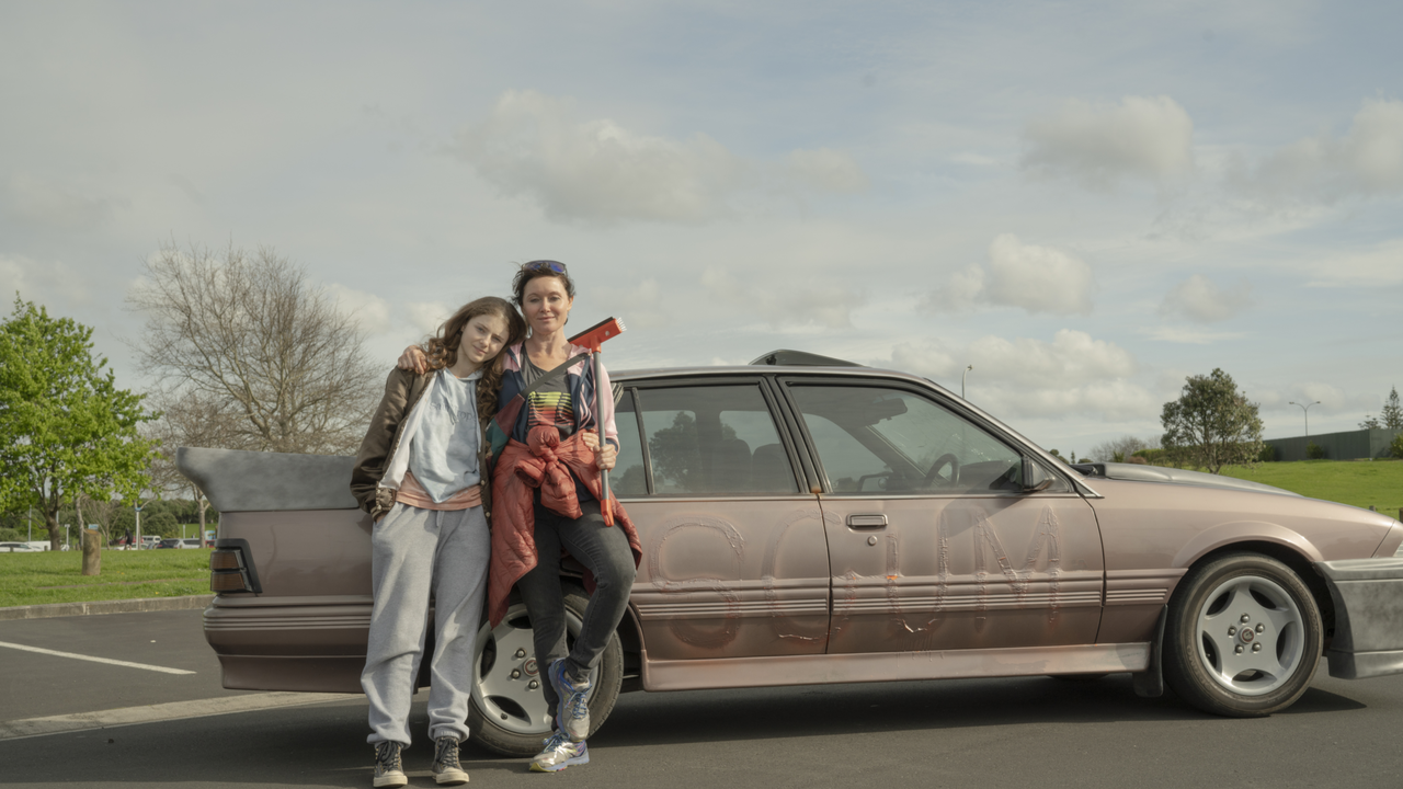 A teenage girl leans against an older woman. A tan car is behind them. They are standing on the road. In the background there is a blue sky with clouds, green grass and trees.