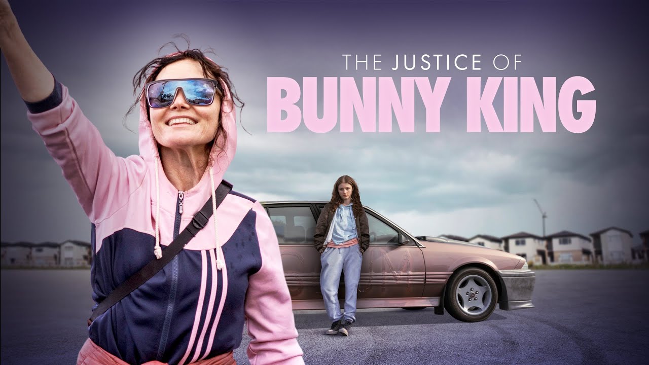 A woman is wearing a pink hoodie. She is wearing sunglasses and is waving at something offscreen. Behind her is an old car and a young teenage girl is leaning aginst it. The Justice of Bunny King title card is light pink.